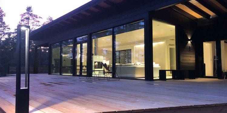 Maison moderne avec de grandes baies vitrées et une spacieuse terrasse en bois, entourée d'arbres. Les intérieurs sont très éclairés et présentent un design minimaliste dans des tons peints à la main. Les lampes d'extérieur LED de LedStore.fi ajoutent une lueur chaleureuse au crépuscule.