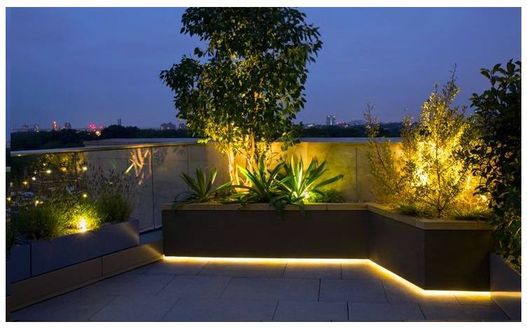 Parterre de fleurs surélevé éclairé par le bas avec une bande de LED aux couleurs chaudes. Le parterre est également équipé d'autres spots aux tons chauds.  