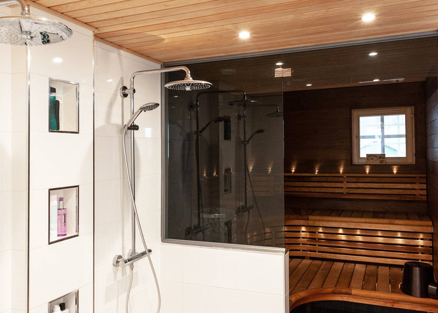 Salle de bain moderne avec deux douches à effet de pluie, paroi en verre, étagère de produits de toilette intégrée, sauna en bois attenant, éclairage d'ambiance par les fenêtres et éclairage LED.