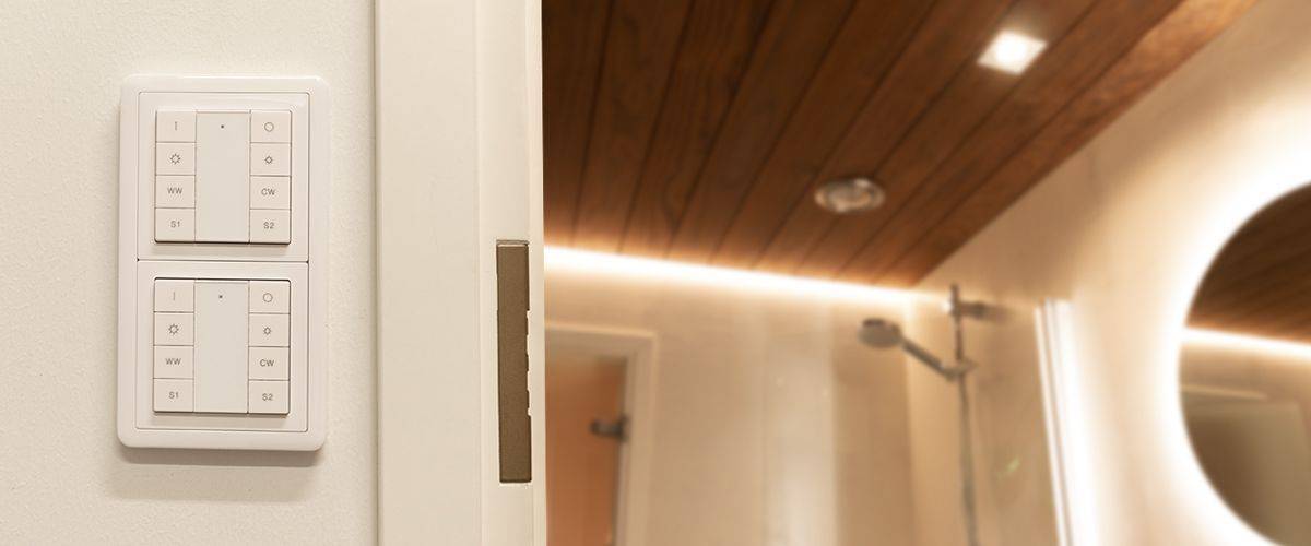 Salle de bains avec panneaux de plafond en bois et éclairage LED doux. Il y a également une douche à l'italienne pratique avec pommeau de douche à effet de pluie. Sur un mur voisin se trouvent deux panneaux d'interrupteurs modernes, commandés par des claviers. L'ensemble est couronné par un miroir circulaire éclairé par des LED.